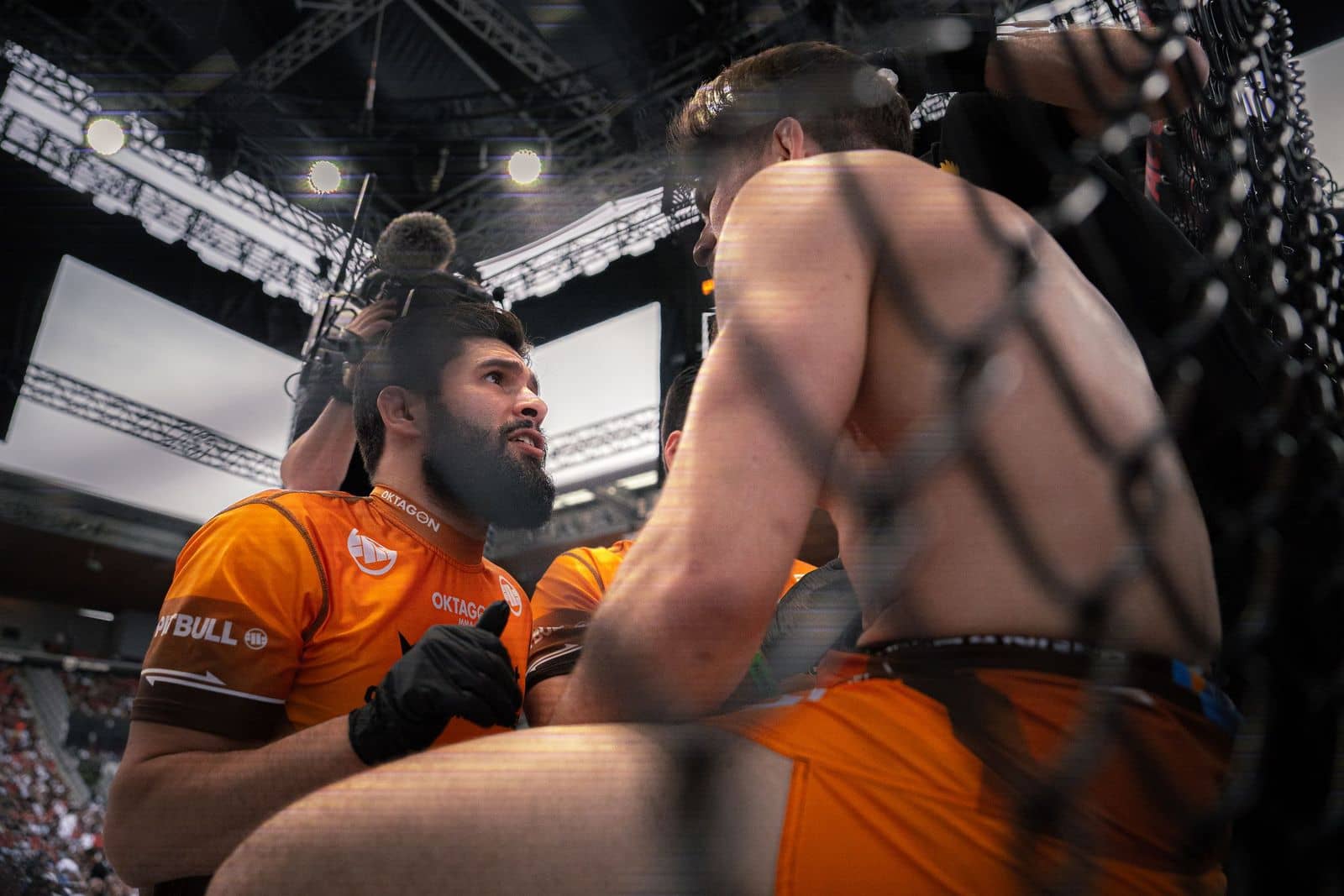 Aus der Betreuerecke nach oben fotografiert: ein Coach im orangefarbenen Team-Trikot blickt zum Käfig hinauf, daneben ein Kameramann.