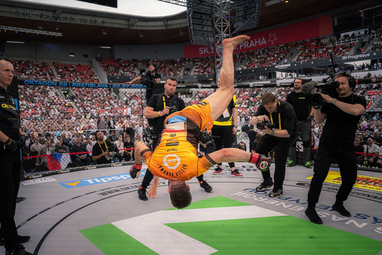Ein Kämpfer schlägt nach dem Sieg einen Salto in der Käfigmitte, rundherum stehen Kameraleute und Fotografen im Fotograben, im Rücken ein voll besetztes Stadion.
